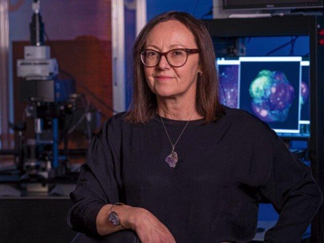 Woman with should length brown hair and glasses wearing a dark shirt standing in a lab