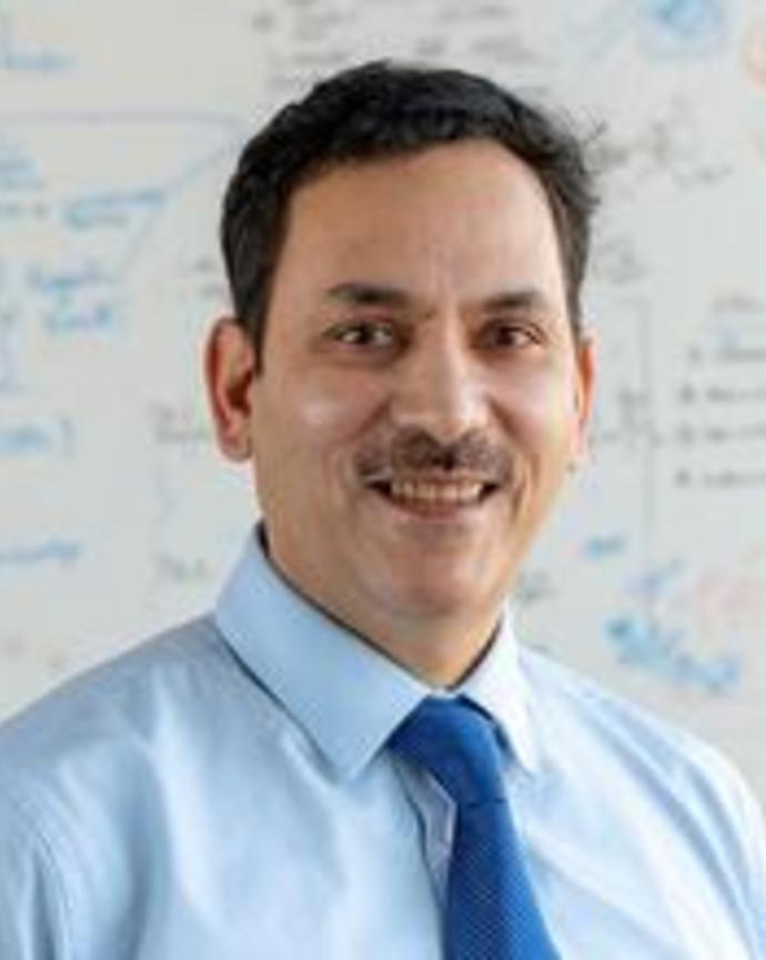 Headshot of man wearing light blue shirt and dark blue tie smiling at camera with a whiteboard in the background