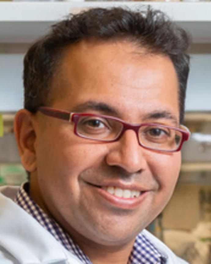 Man with short dark hair and red glasses wearing a lab coat smiling in front of blurred lab setting